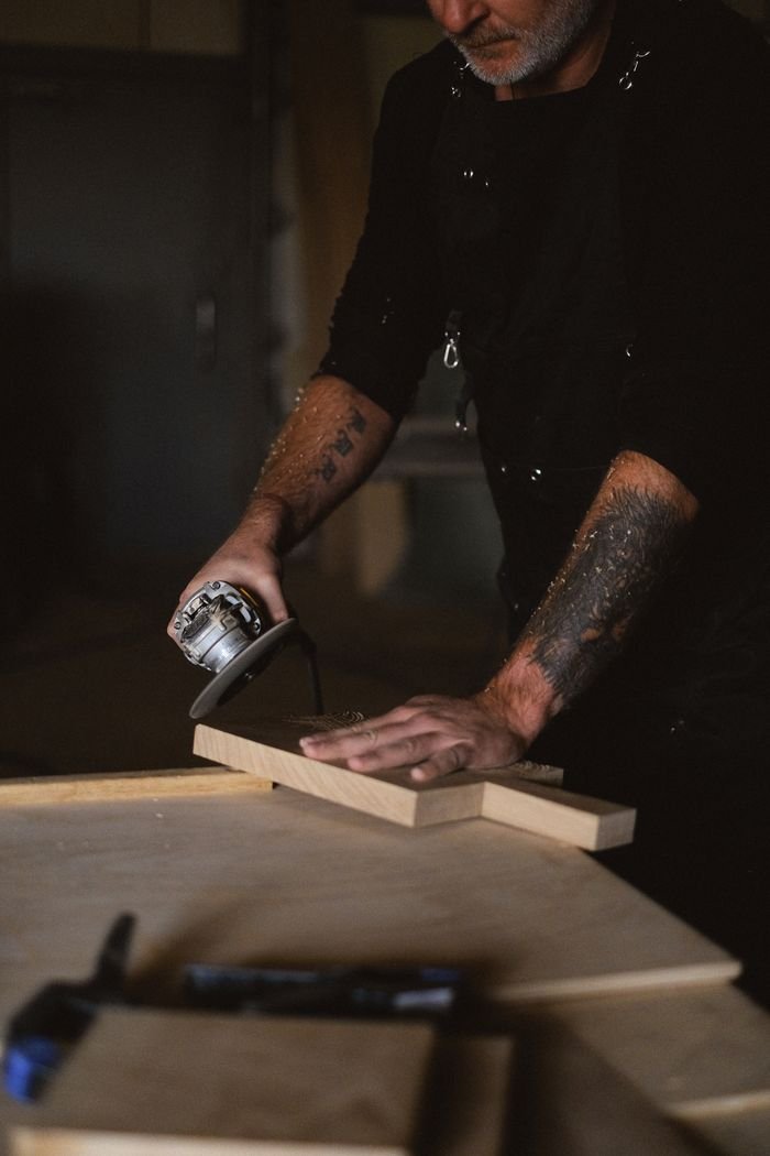 Unrecognizable tattooed male artisan with angle grinder polishing wooden board while standing at table in professional joinery with special equipment