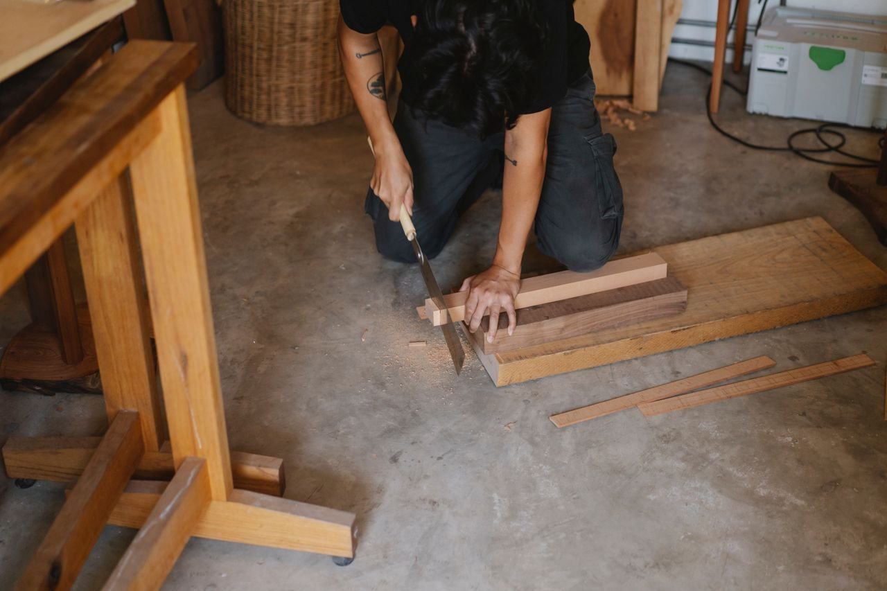 Anonymous joiner working with Japanese saw while cutting edge of timber plank on floor in workshop